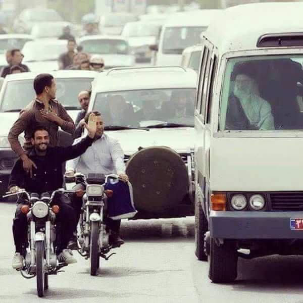 Khamenei travelling in an ordinary van among the civilian traffic, two men on a motorbike waving to him